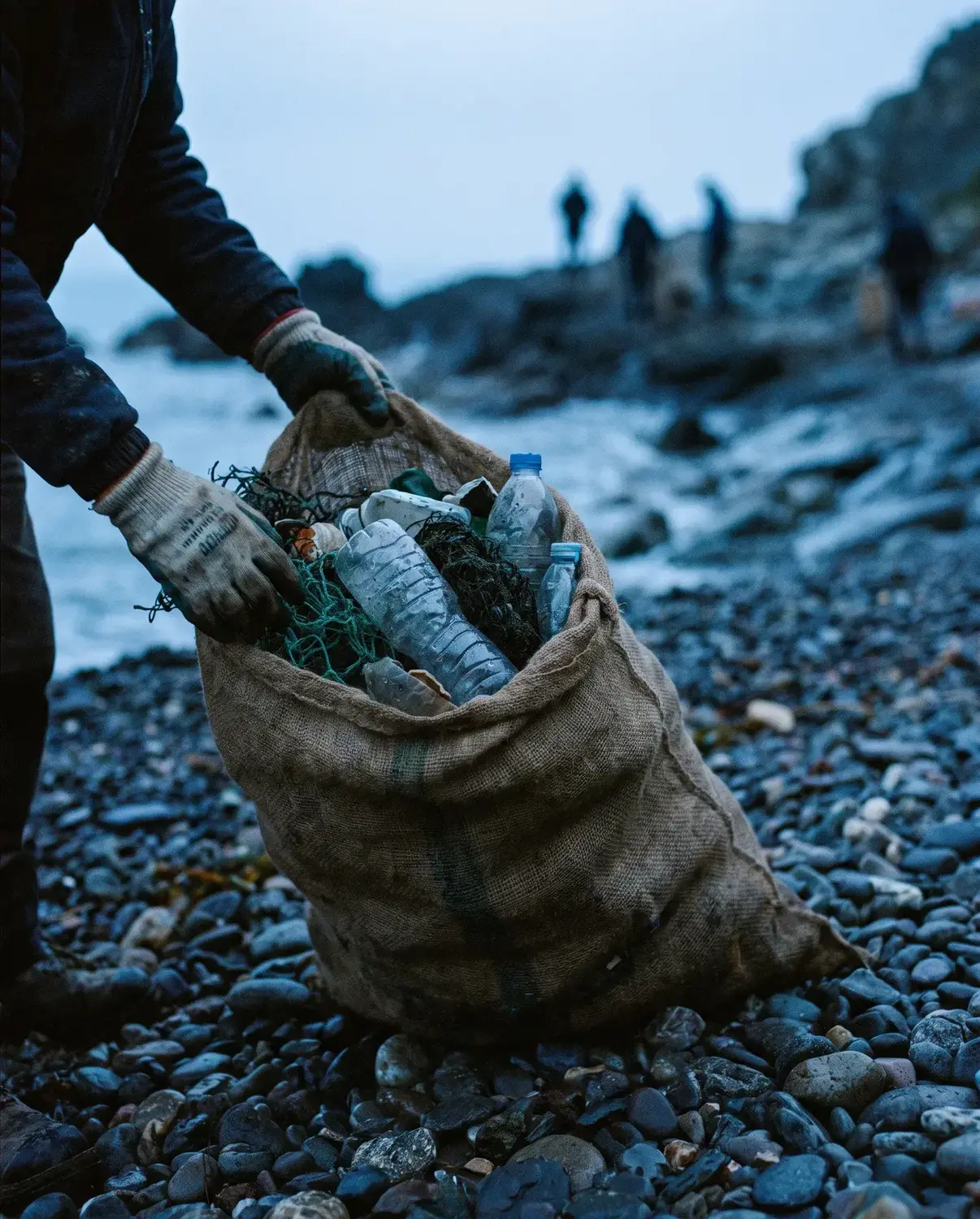 Gloved hands lifting a sack of collected waste on a stony shore
