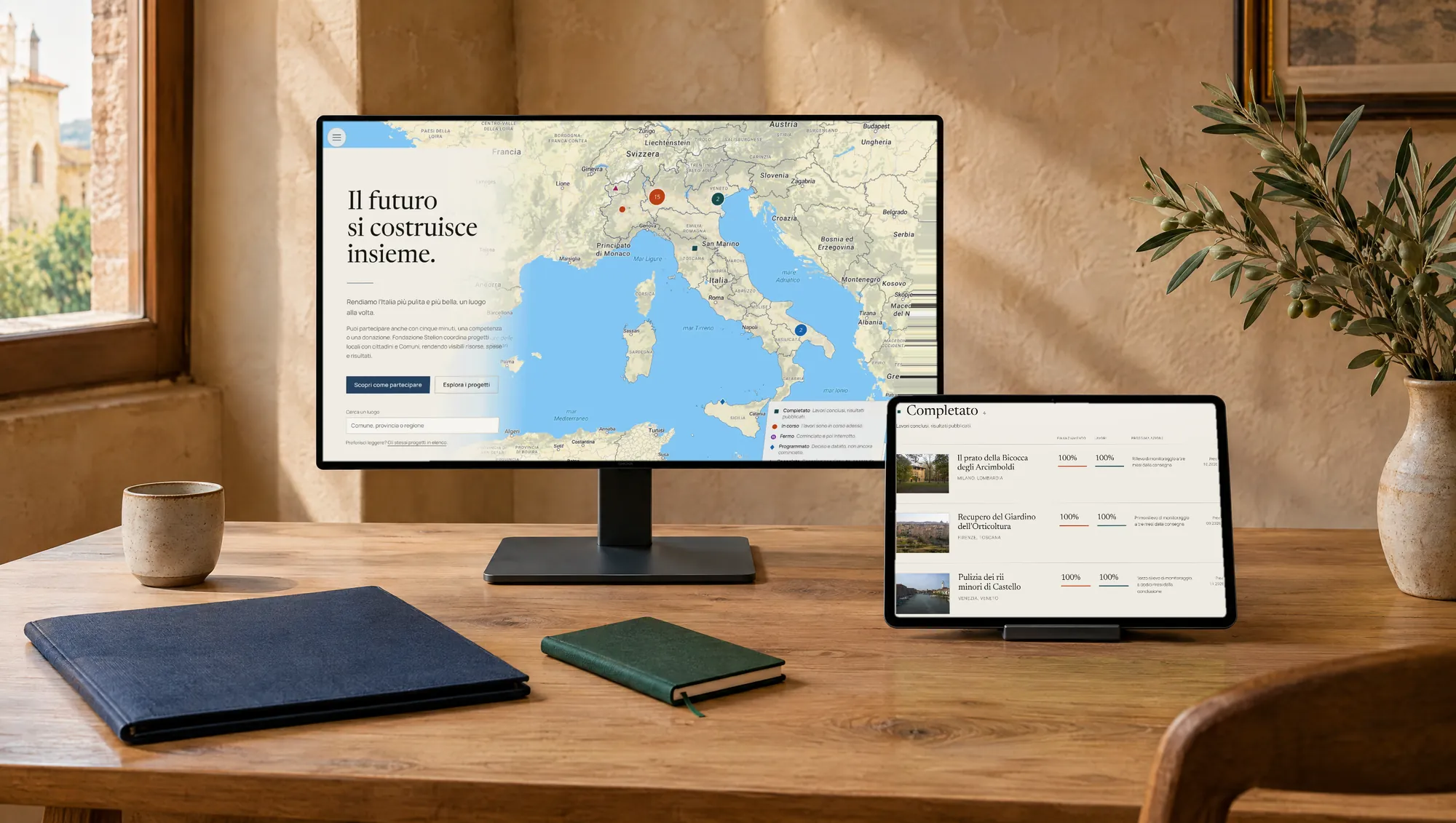 The Fondazione Stelion website on a monitor and a tablet, on a wooden table in a warm-walled room
